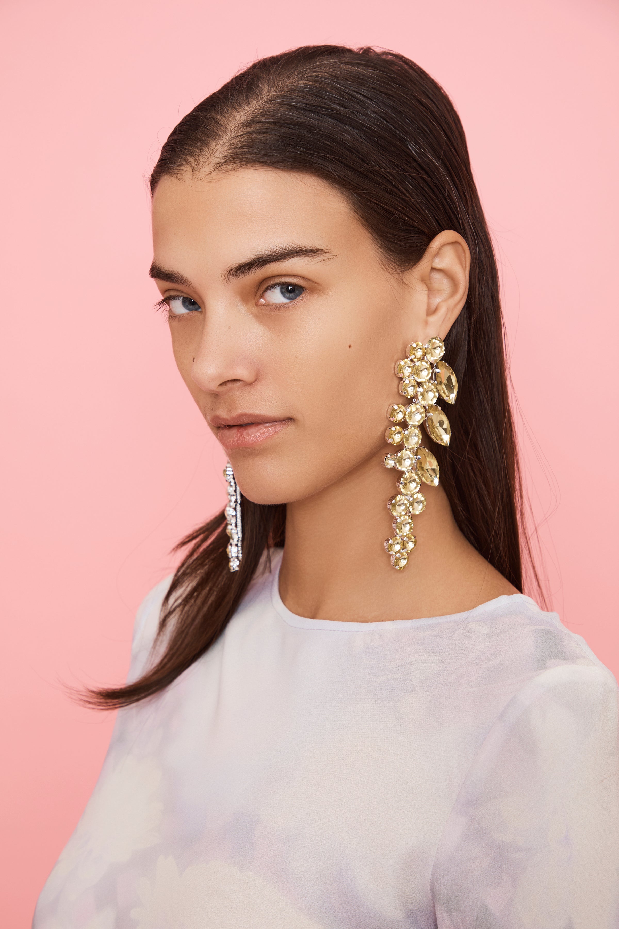 Woman wearing large draping floral-shaped yellow rhinestone earrings against a pink background
