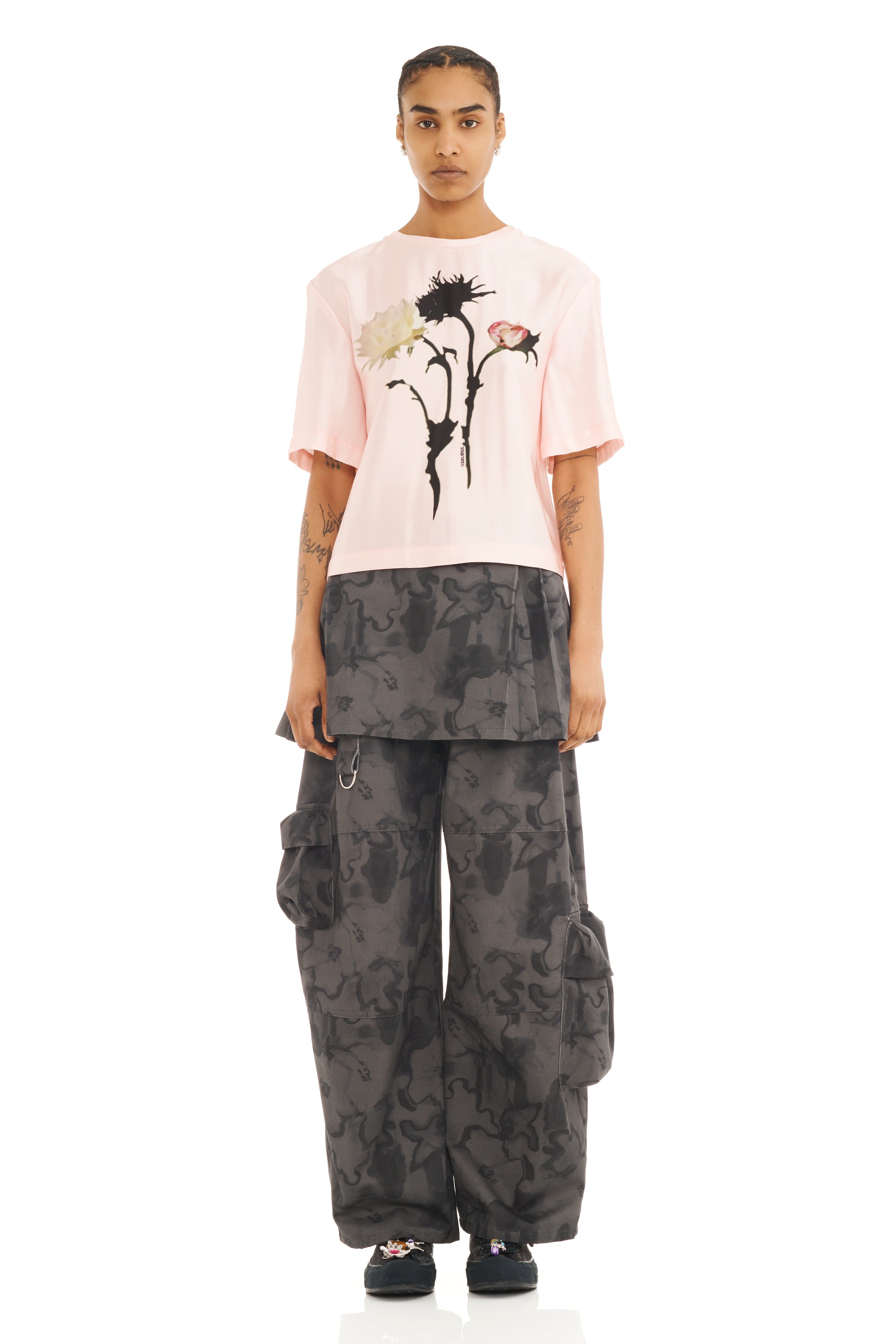 Person wearing a pink t-shirt with elbow length sleeves and a floral graphic design and camouflage pants on a white background