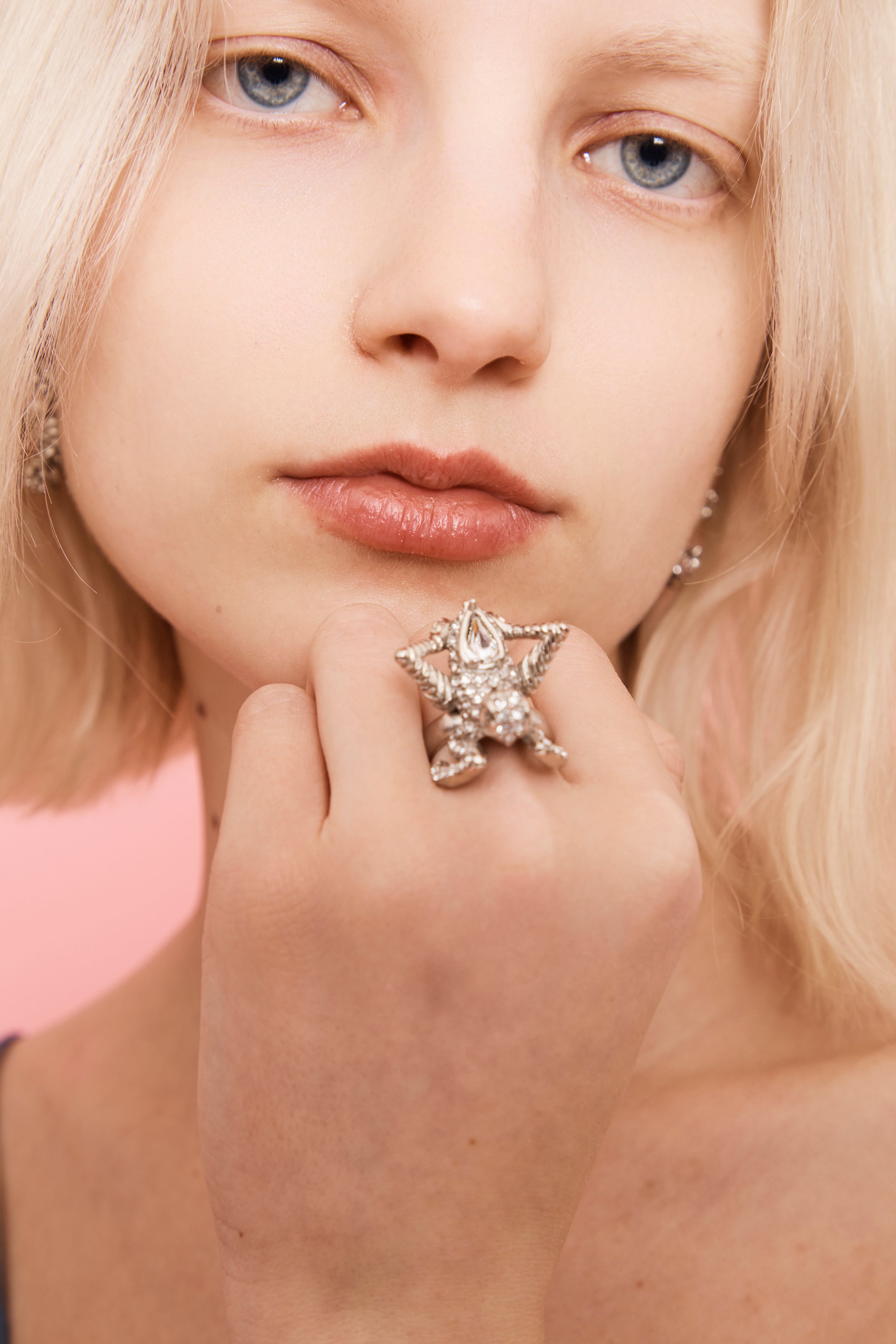 Person wearing a praying mantis-shaped ring encrusted with clear rhinestones on a pink background