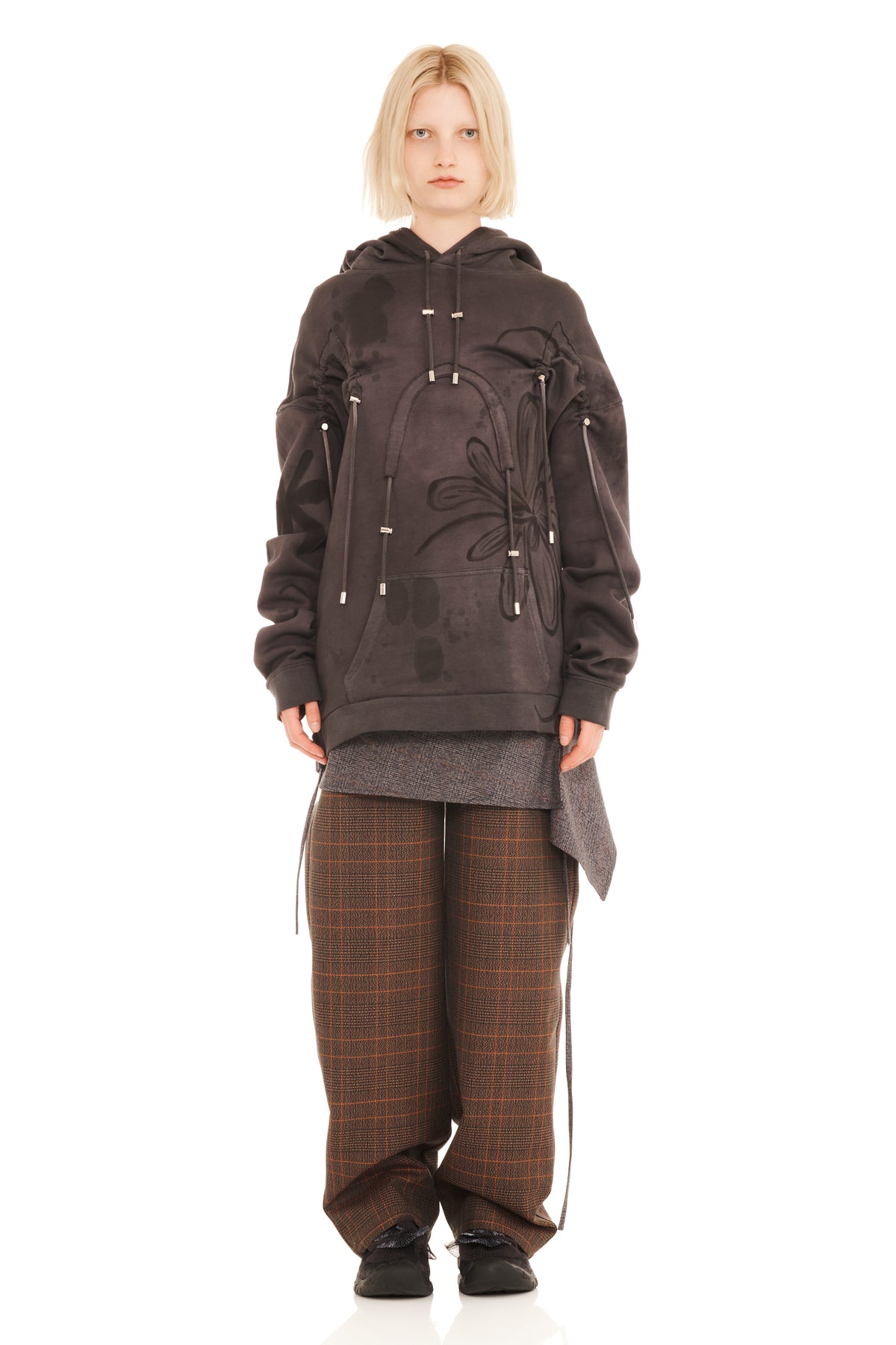A model stands against a white studio background wearing a dark brown hooded sweatshirt with tonal floral embroidery and silver drawstring details, layered over a gray shirt, paired with brown plaid trousers and black shoes.