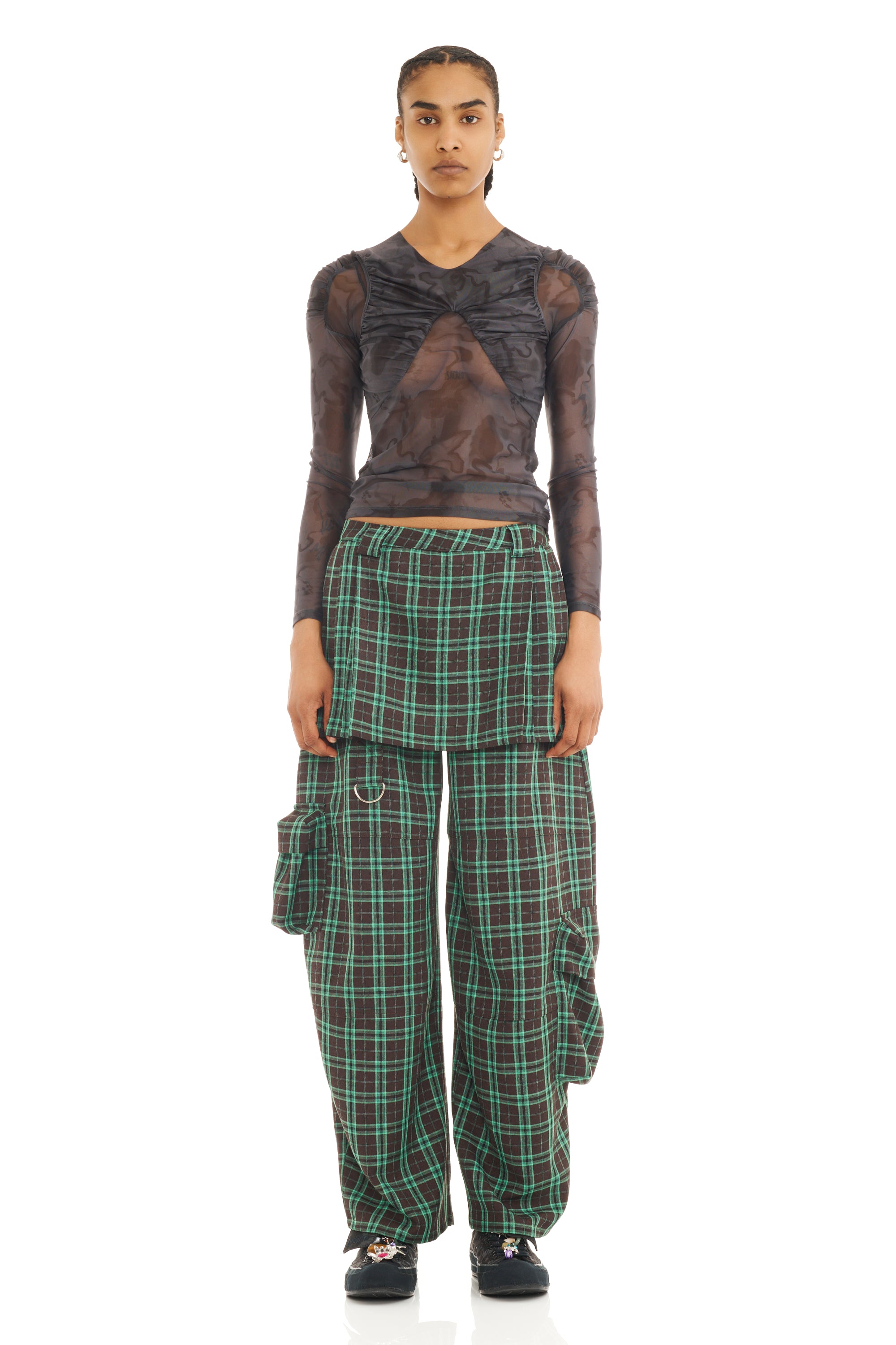 Woman wearing a sheer black top with a green and brown plaid pant with matching skirt overlay on a white background.